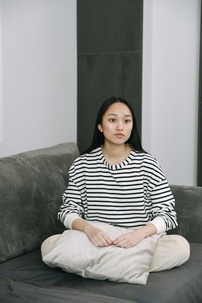 A young woman in a striped shirt sits on a couch indoors, looking contemplative.