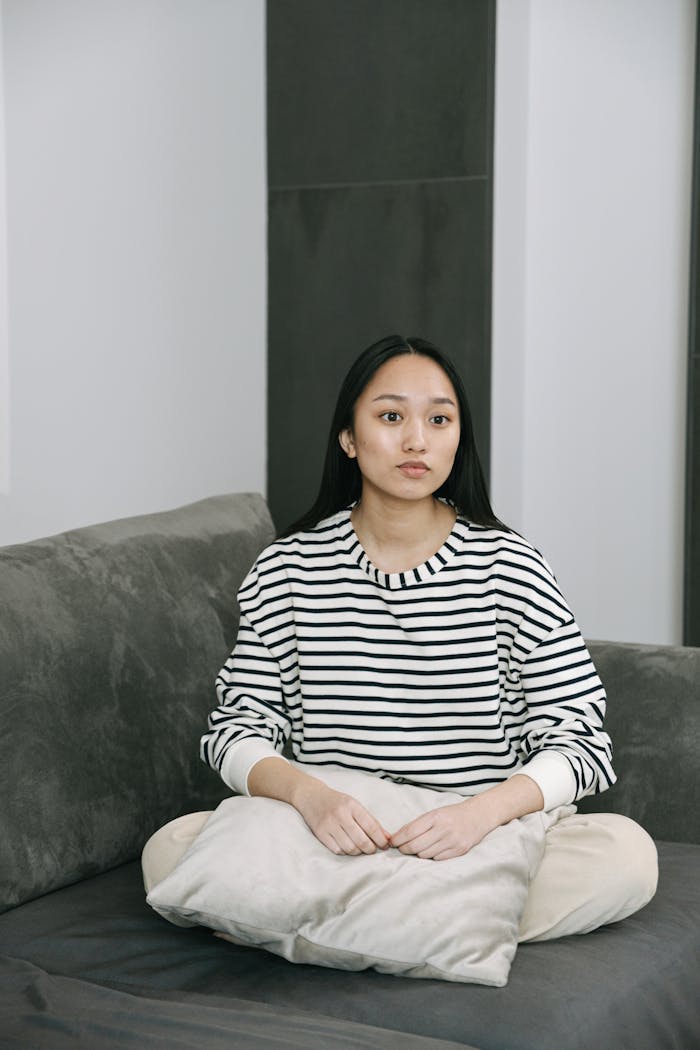 A young woman in a striped shirt sits on a couch indoors, looking contemplative.