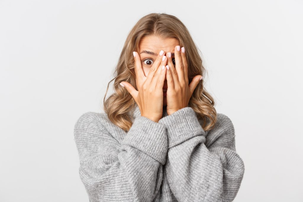 close up of shocked or embarrassed blond girl, cover eyes and peeking through fingers, standing over white background